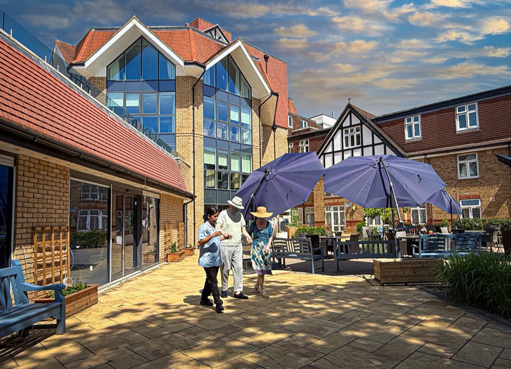 A carer walking with two residents across the sunny courtyard garden at Forest Place Nursing Home, Buckhurst Hill
